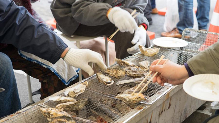 A group of people are preparing oysters on a grill for the Hinase Kaki Matsuri Oyster Festival