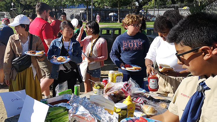 People of diverse ages and backgrounds gathering around a table with food at an outdoor community event.