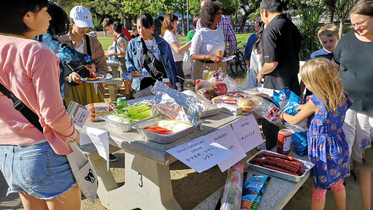 People gathered around a picnic table with various dishes and ingredients during an outdoor potluck event.