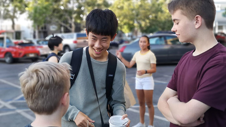 A group of teenagers conversing in a parking lot, with one smiling asian boy holding a backpack in the center.