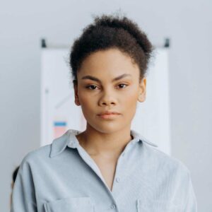 A woman with short curly hair wearing a light blue button-up shirt stands indoors, looking directly at the camera with a neutral expression—perfect for an "about our company" introduction.