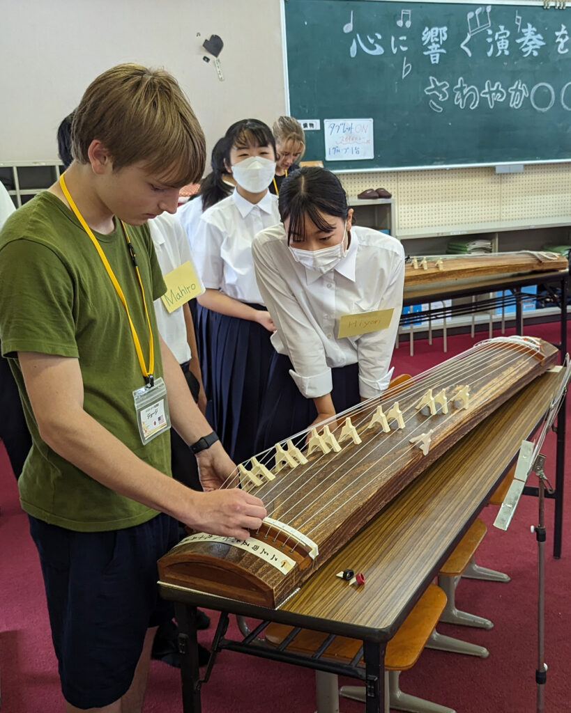In Japan, a young man learns to play a koto, observed by students in uniforms as part of a cultural exchange. A blackboard with writing is seen in the background.