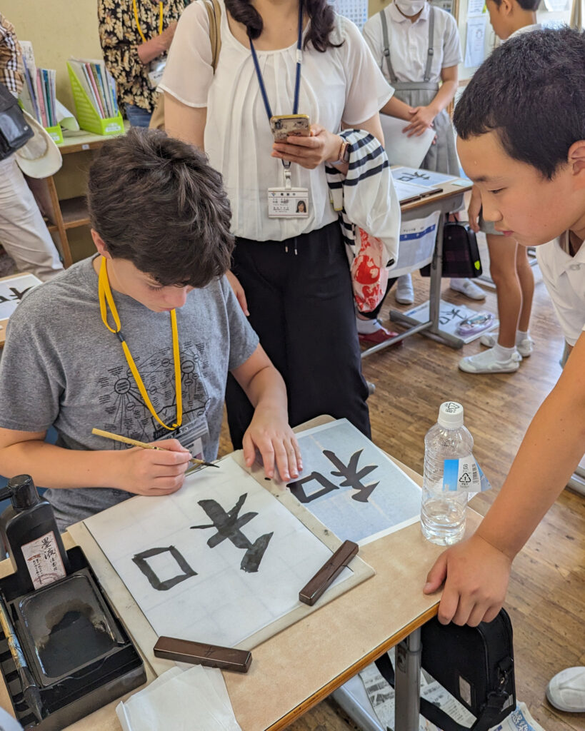In a classroom during a Japan student cultural exchange, students practice calligraphy. One scribes on paper while another observes intently. The teacher and classmates stand nearby, with bottled water and a Bizen ink stone on the desk, reflecting the essence of Japanese tradition.