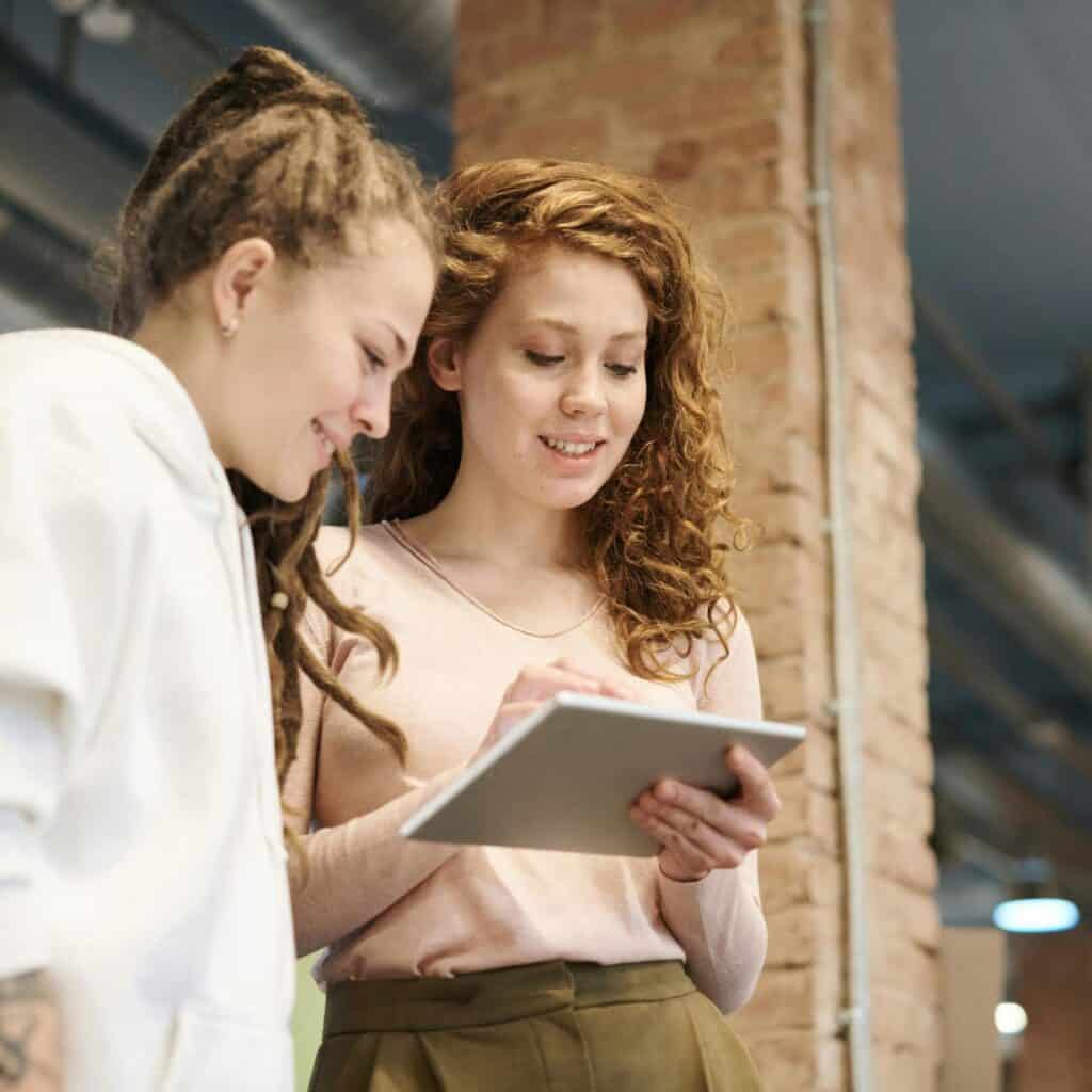 Two women standing indoors, smiling and looking at a tablet device together, with exposed brick and industrial-style decor in the background.
