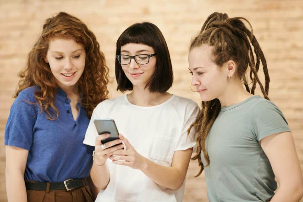Three young women stand together, looking at a smartphone one of them is holding, all appearing engaged and interested.