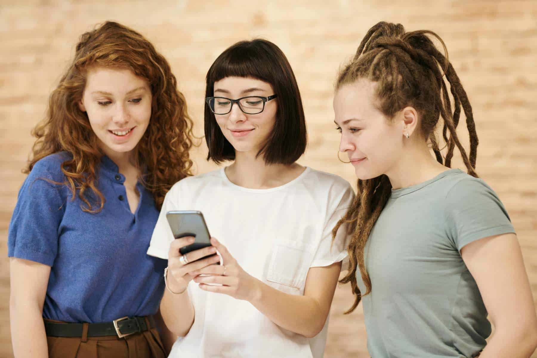 Three young women stand together, looking at a smartphone one of them is holding, all appearing engaged and interested.