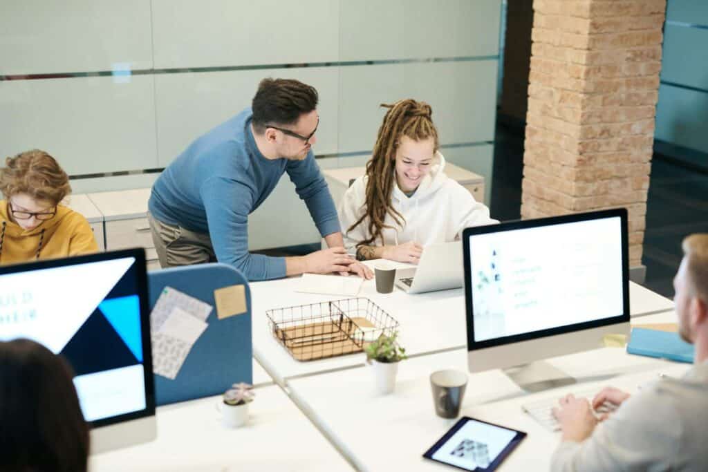 Four people work at desks with computers in a modern office; one person stands and speaks with a colleague who is smiling and using a laptop.