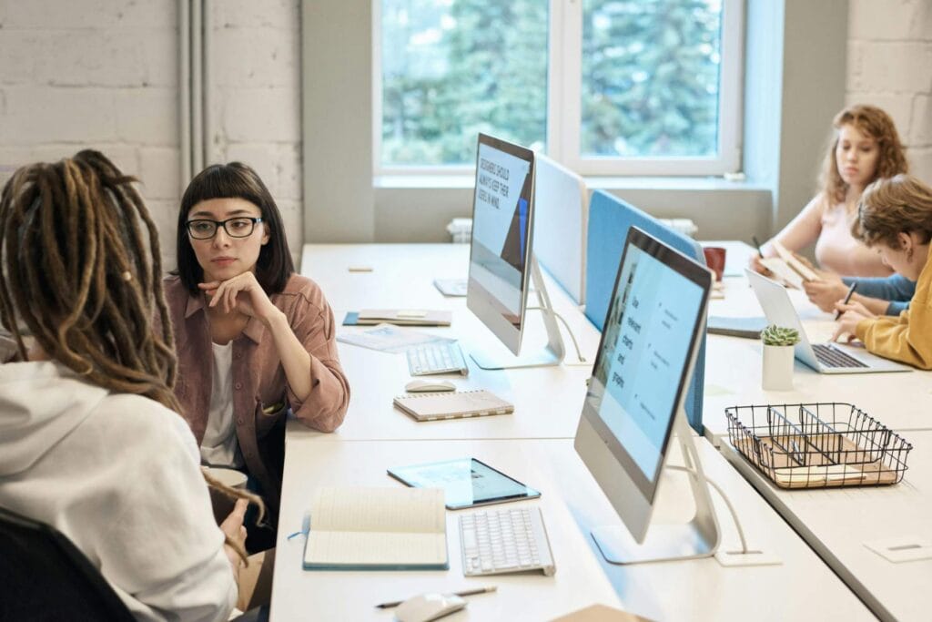 Four people work at a shared desk with computers in a modern office. Two are in conversation while two others focus on their laptops and notebooks.