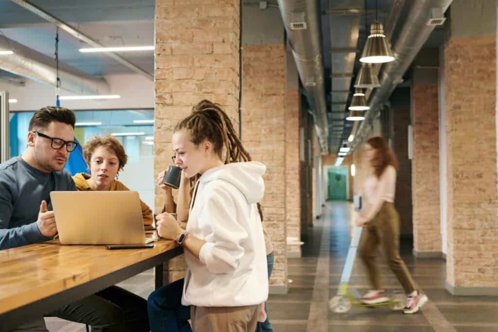 Four people sit at a high table working on a laptop in a modern office space; another person rides a scooter down the hallway in the background.