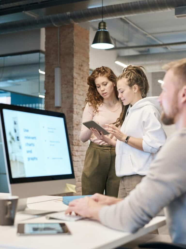 Two women discuss something on a tablet while a man works at a computer with charts displayed on the screen in a modern office setting.