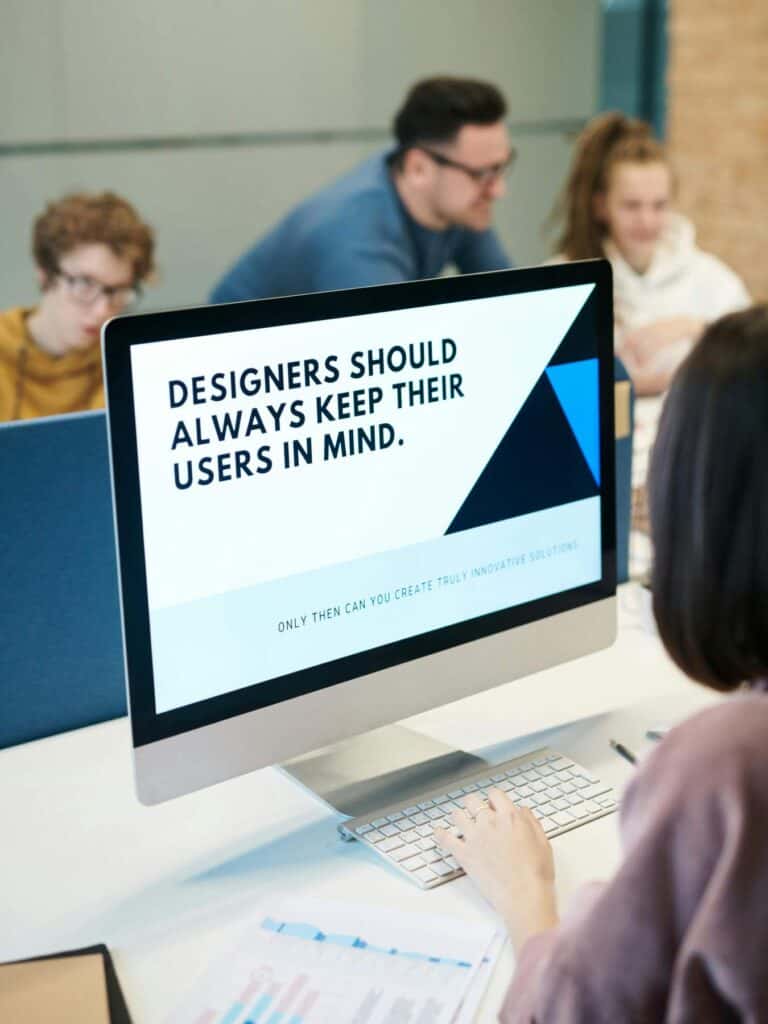A computer screen displays the message "Designers should always keep their users in mind," while people work together at a table in the background.