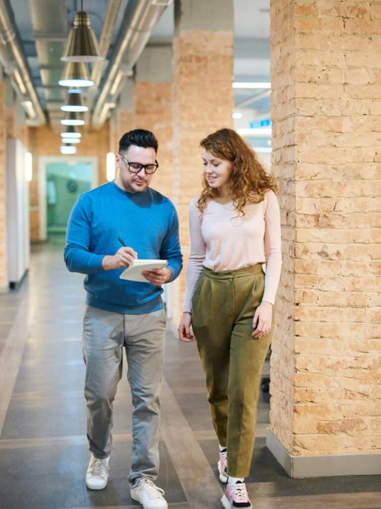 Two people walk down a hallway with exposed brick walls; the man writes on a notepad while the woman walks beside him, both engaged in conversation.