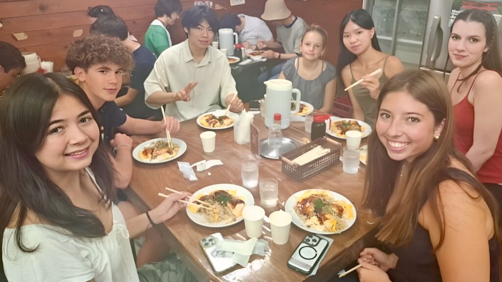 A group of young people sit around a wooden table in a restaurant, enjoying meals with chopsticks and smiling at the camera—a joyful moment of cultural exchange between Menlo Park Sister Cities and Bizen, Japan.