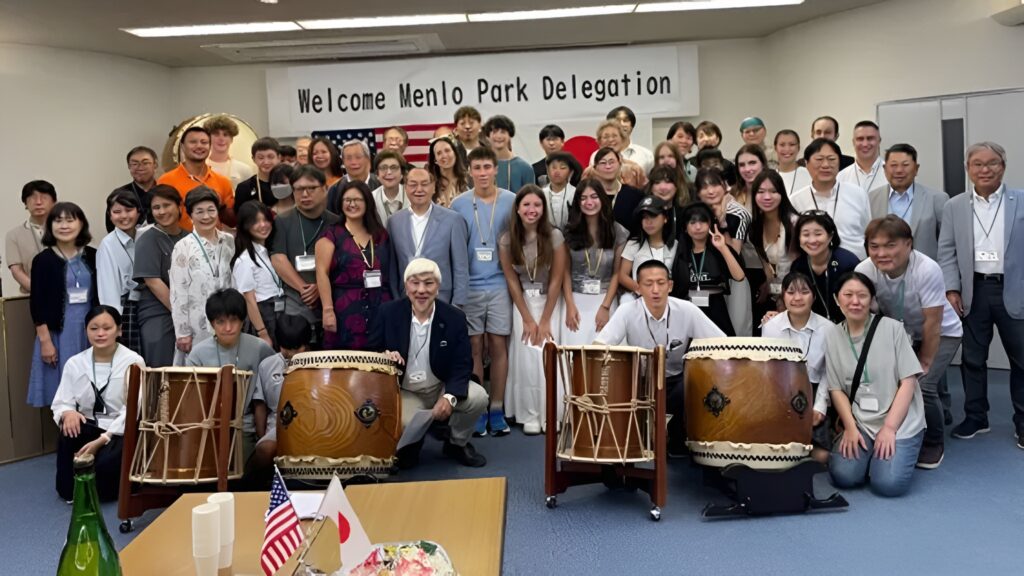 A large group of people pose together indoors under a banner reading "Welcome Menlo Park Delegation," celebrating the Menlo Park Sister Cities experience with traditional Japanese drums and US and Japanese flags in front, highlighting Bizen Japan.