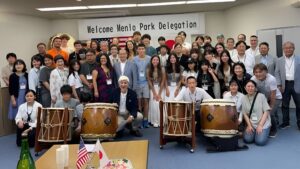 A large group of people pose together indoors under a banner reading "Welcome Menlo Park Delegation," celebrating the Menlo Park Sister Cities experience with traditional Japanese drums and US and Japanese flags in front, highlighting Bizen Japan.