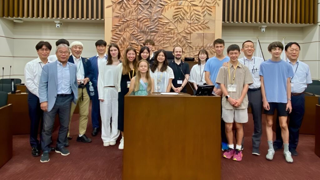 A group of adults and teenagers from Menlo Park Sister Cities pose together in a conference room with a large decorative wooden panel in the background, sharing their My Experience visit to Bizen, Japan.