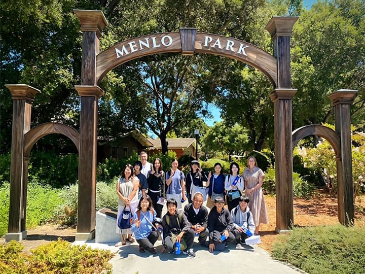 A group of people, including host families, pose for a photo under a large wooden archway labeled "Menlo Park," surrounded by trees and greenery on a bright summer 2026 day.