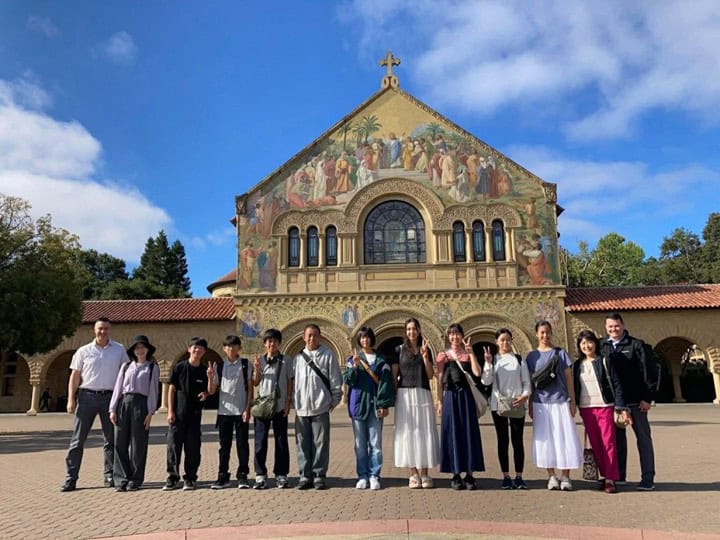 A group of people, including host families, pose for a photo in front of a historic building with a colorful mural and arched windows on a sunny summer 2026 day.