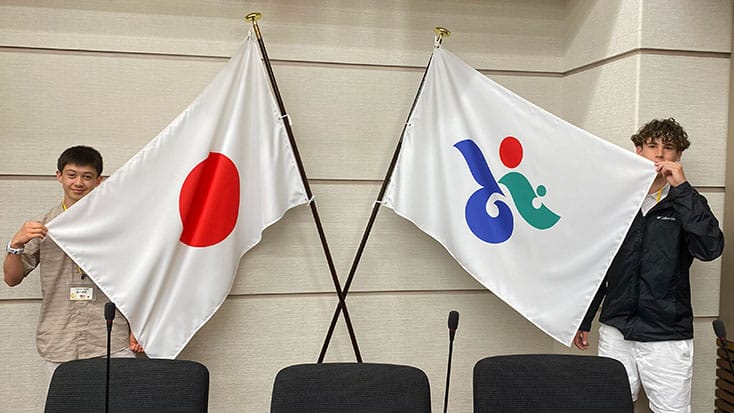Two young men from a Bizen delegation stand indoors holding flags; one is Japan's national flag, the other is a white flag with a blue, green, and red abstract symbol—perhaps representing their Menlo Park visit.