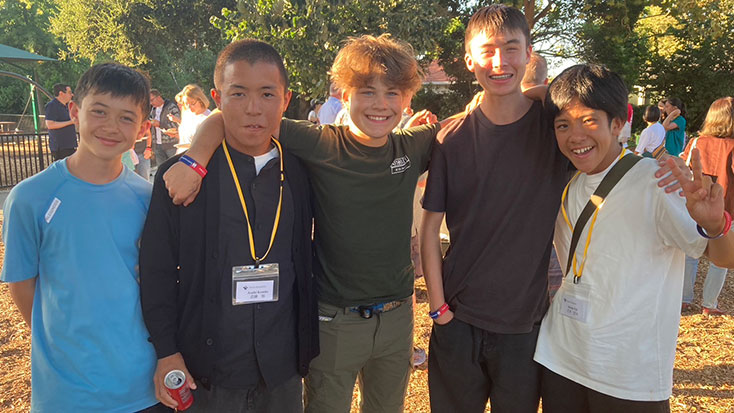 Five teenage boys, including Japanese students, stand outdoors in Menlo Park smiling at the camera. One holds a drink can. Other people and trees in the background suggest they're hosting or attending a lively social gathering.