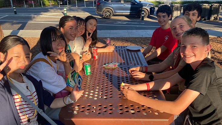 A group of eight teenagers sit around a metal picnic table outdoors in Menlo Park, playing Uno and drinking soda while hosting Japanese students, with cars parked in the background.