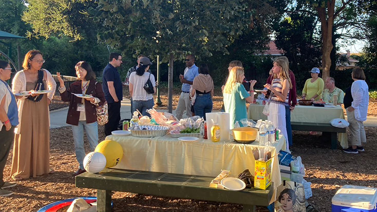 A group of people socialize and gather food around picnic tables covered with snacks and drinks in Menlo Park, hosting Japanese students in a sunny park setting.