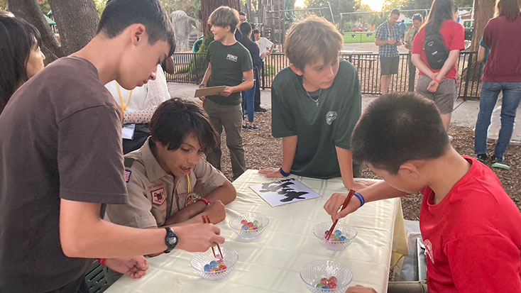 Several boys, including Japanese students, gather around a table outdoors in Menlo Park, using chopsticks to pick up colorful objects from bowls while others observe or stand nearby.