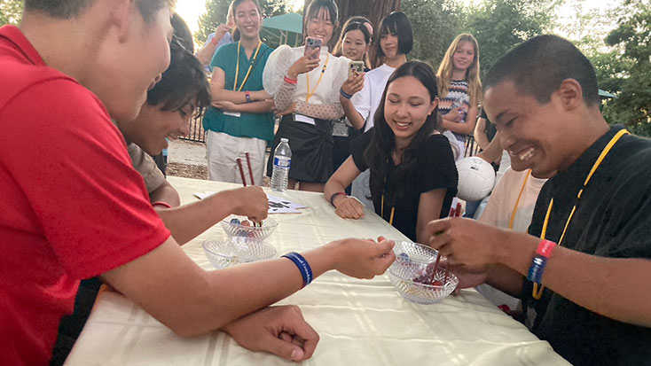 Four Japanese students sit at a table in Menlo Park, participating in a contest with bowls, while a group of onlookers stands behind them, watching and smiling.
