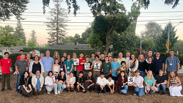 A large group of people, including adults and children, pose outdoors in Menlo Park in front of trees and houses. Some individuals, possibly hosting Japanese students, hold up papers with Japanese calligraphy.