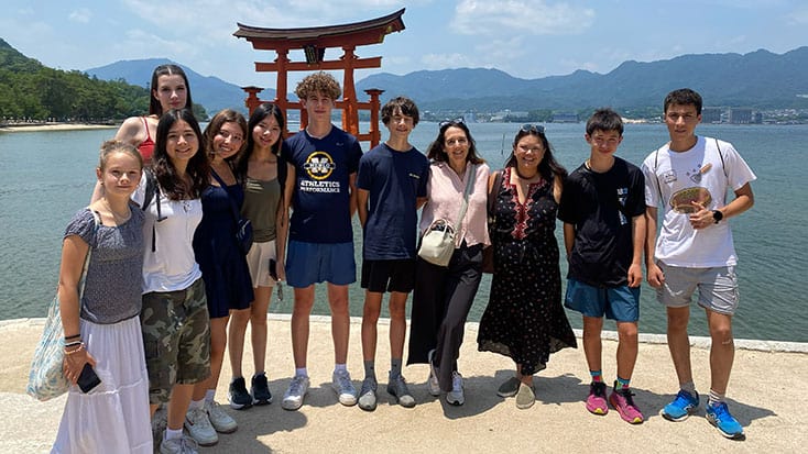 A group of ten young people, part of a delegation from Menlo Park, pose in front of the floating torii gate at Itsukushima Shrine on a sunny day, with mountains and water in the background.