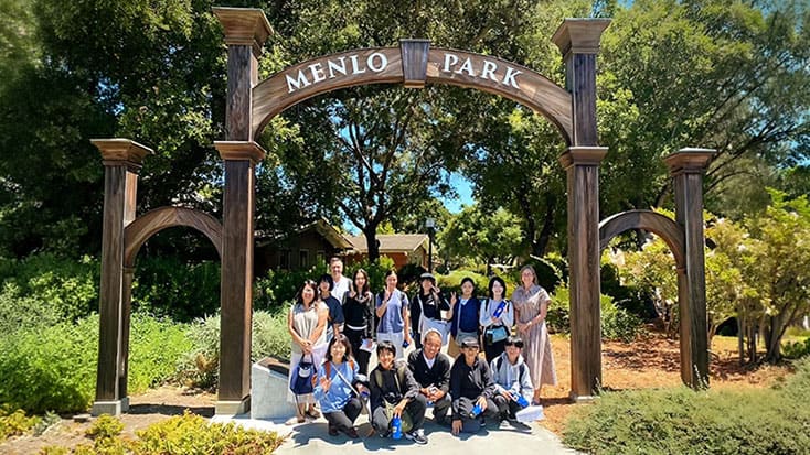 A group of people pose for a photo under a large wooden archway labeled "Menlo Park," surrounded by greenery and trees.